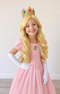 Child modeling a blonde wig, crown headband, white gloves, earrings, and blue brooch with a pink dress